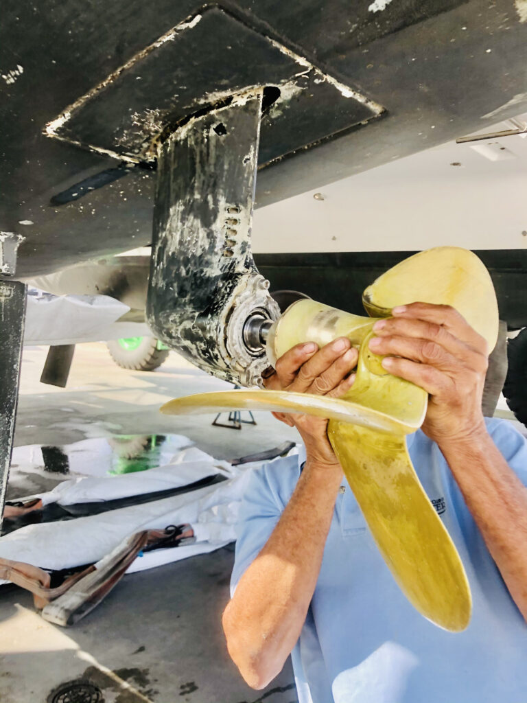 Hands seating a bronze three-blade prop on a saildrive under a hauled-out hull